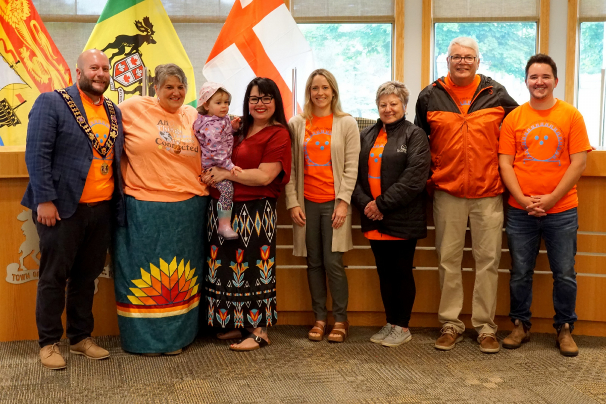Riverview town council members and staff and indigenous community members gather in front of mikmaq grand council flag displayed in council chambers