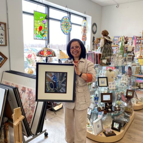 Interior of Kay's Custom Framing. Shop owner (Connie) holding a picture frame with artwork. The background shows shop frames, giftware and artwork.