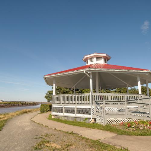 Riverfront gazebo with a white ramp and railings. It has a red roof. The background shows a gravel path, flowers, grass and the river.