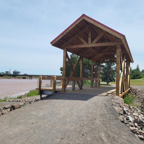 Front view of gravel path leading up to the wooden bridge with a red roof. Background shows stones, grass and the river.
