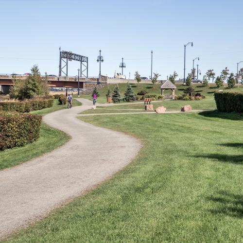 View of the riverfront trail. Shows a s shaped gravel path trailing off in the distance. The background shows 2 cyclists, the third wave stone sculpture, the gunningsville bridge and some grass/greenery.