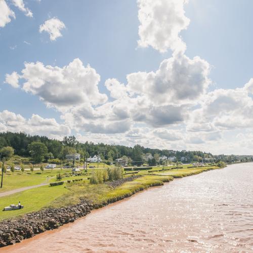 Shows the riverfront at a distance. The top half of the image is a light blue sky with fluffy white clouds. The bottom right corner shows the riverfront trail and surrounding greenery. The right bottom corner shows the chocolate river.