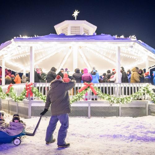A gazebo in Christmas lights with a man pulling two kids bundled up in a wagon 