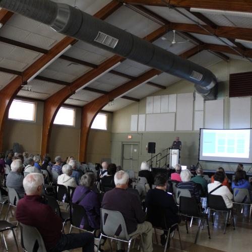 A speaker with powerpoint screen presenting to a crowd of people in chairs