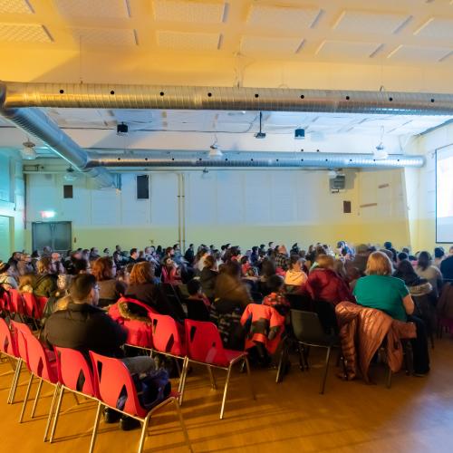 A crowd in chairs watching a projector in a community hall