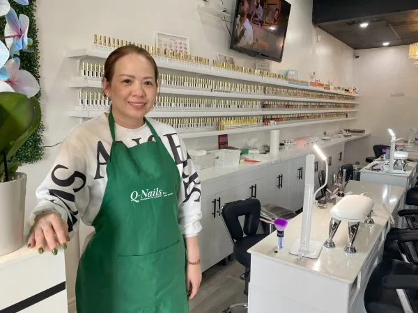 A woman in a green apron standing in her nail salon, with hundreds of nail polish bottles on shelves in the background