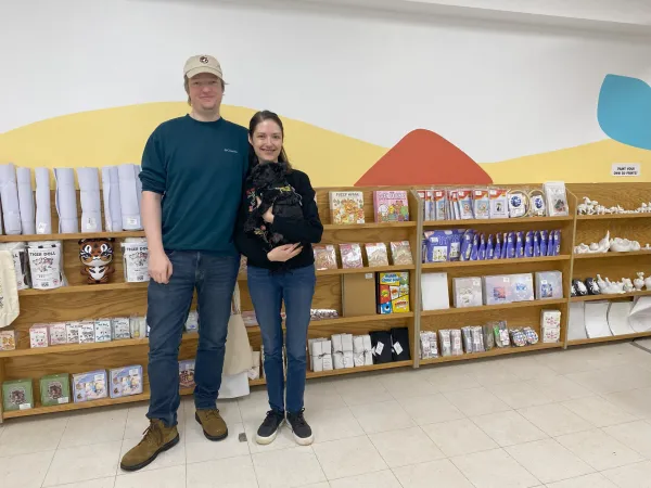 A couple smiling with their dog in front of shelves of art projects and supplies