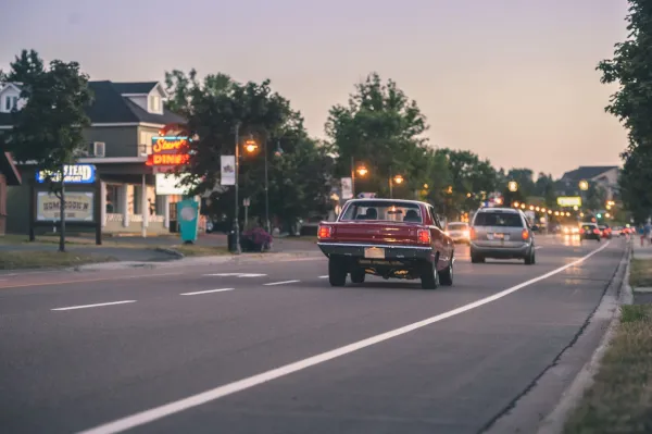 A red car driving down Coverdale Road as the sun sets. The Homestead restaurant, Steeve's dinner, trees and streetlights are visible in the background.