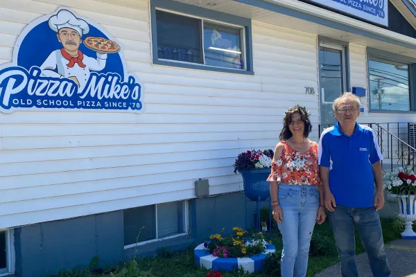 A man and woman smiling in front of white building with blue sign