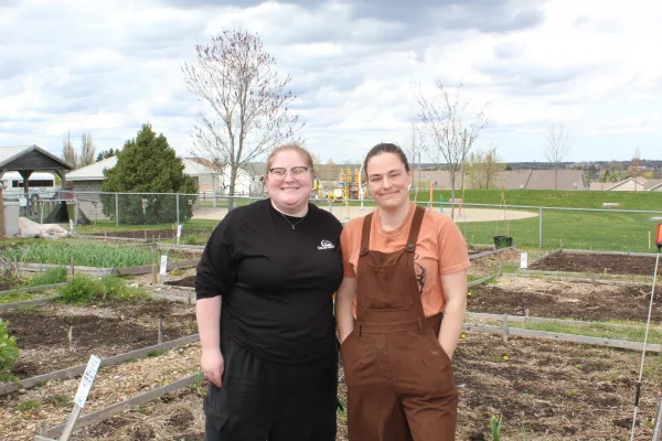 Kass (left) and Alicia (right) stand together in the Coverdale Community Garden, surrounded by garden beds and greenery. Both are smiling as they pose outdoors in the community garden space.
