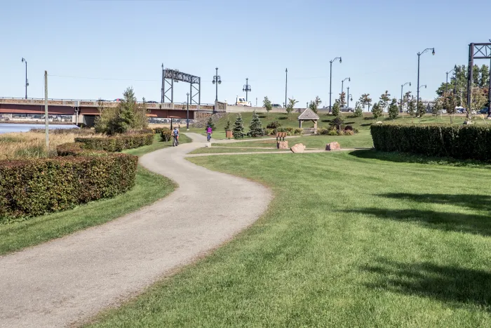 View of the riverfront trail. Shows a s shaped gravel path trailing off in the distance. The background shows 2 cyclists, the third wave stone sculpture, the gunningsville bridge and some grass/greenery.