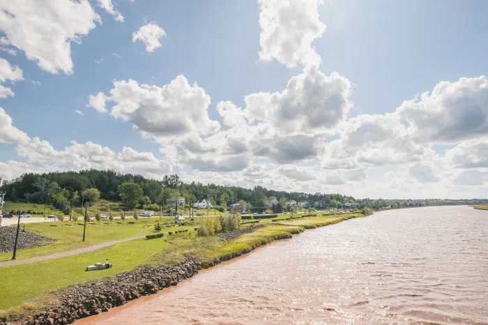 Shows the riverfront at a distance. The top half of the image is a light blue sky with fluffy white clouds. The bottom right corner shows the riverfront trail and surrounding greenery. The right bottom corner shows the chocolate river.