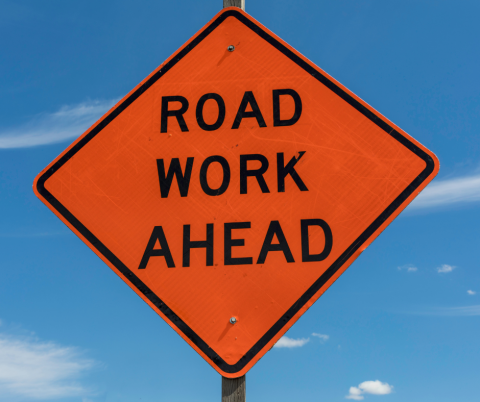 Orange diamond sign says road work ahead against blue sky