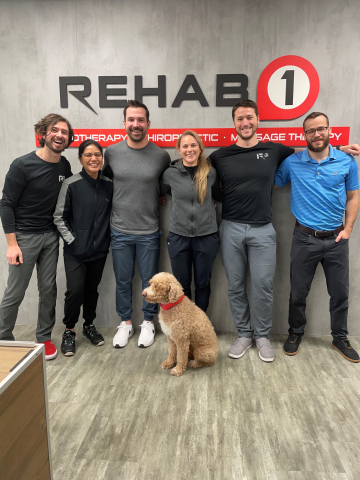 six team members and a golden doodle stand in front of Rehab 1 sign in office