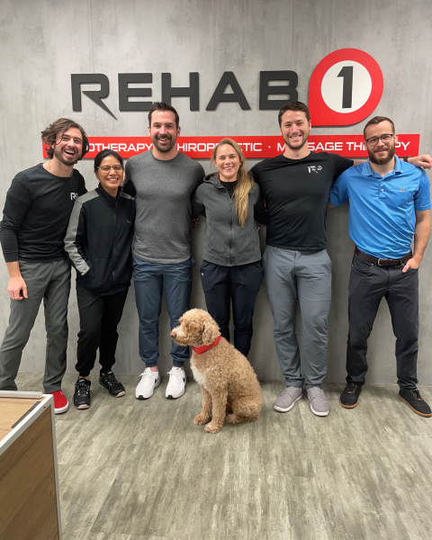 six team members and a golden doodle stand in front of Rehab 1 sign in office