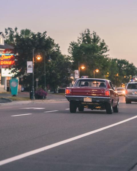 A red car driving down Coverdale Road as the sun sets. The Homestead restaurant, Steeve's dinner, trees and streetlights are visible in the background.