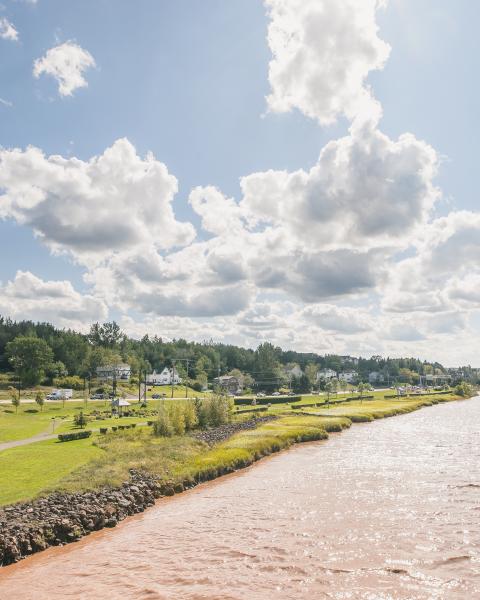 Shows the riverfront at a distance. The top half of the image is a light blue sky with fluffy white clouds. The bottom right corner shows the riverfront trail and surrounding greenery. The right bottom corner shows the chocolate river.