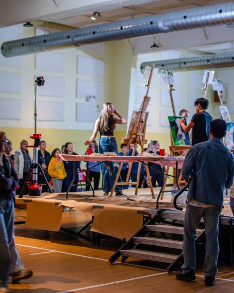 Youth participating in a live painting competition surrounded by an audience. 
