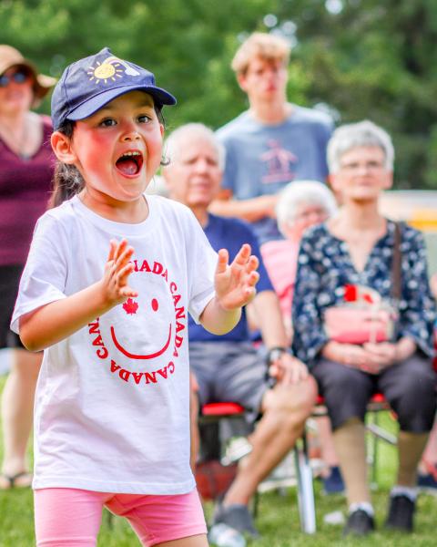 A young girl wearing a blue ball cap and a white Canada t-shirt clapping happily.