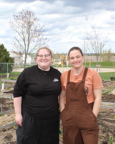 Kass (left) and Alicia (right) stand together in the Coverdale Community Garden, surrounded by garden beds and greenery. Both are smiling as they pose outdoors in the community garden space.