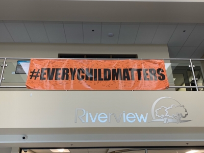 Orange vinyl banner that reads 'Every Child Matters' hangs on the mezzanine in Riverview Town Hall. It is marked with dozens of black signature. 