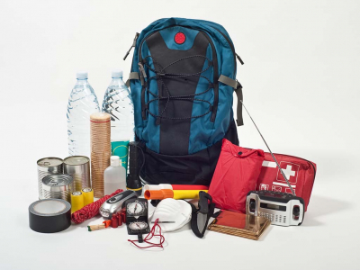 An emergency preparedness kit is shown including a large backpack surrounded by items such as water bottles, tape, face masks, canned food, a flashlight, flares, warming blanket, radio, etc. 